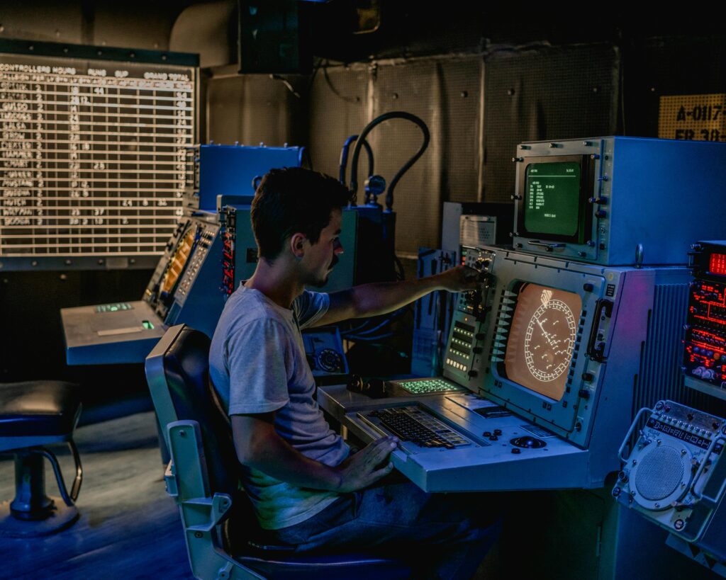 A man working at control panels in a dimly lit industrial room with various monitors.