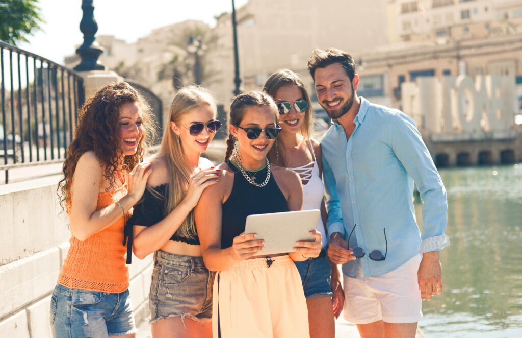 Five friends laughing together by the waterfront in Malta, enjoying a sunny day and sharing a tablet.