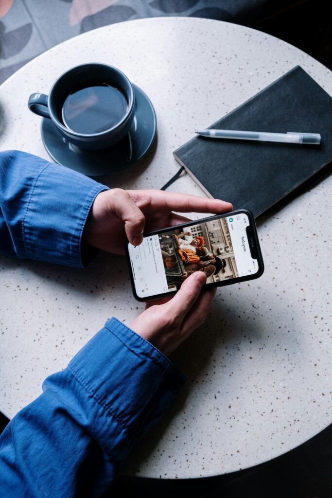 A person views Instagram on a smartphone at a cafe table with coffee, notebook, and pen.